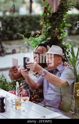 Un couple de personnes âgées souriant prend le selfie sur un téléphone intelligent tout en étant assis dans le café-terrasse Banque D'Images