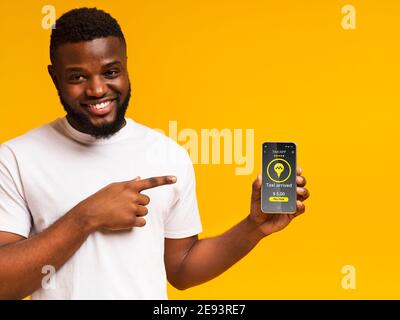 Joyeux homme noir pointant vers un smartphone avec une interface d'application de taxi, commandant un taxi, utilisant le service mobile sur fond orange Banque D'Images