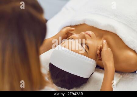 Cosmetologiste faisant le soin de massage facial pour la jeune femme sereine dans salon de beauté Banque D'Images