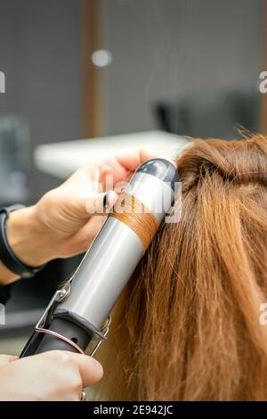 Coiffeur professionnel fait des boucles avec un fer à friser pour un jeune femme aux longs cheveux rouges dans un salon de beauté Banque D'Images