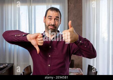 Jeune beau barbu homme portant une chemise mauve faisant des pouces vers le haut et vers le bas, en désaccord et d'accord expression. Concept de conflit Banque D'Images