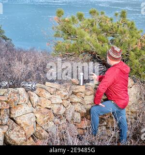 un randonneur senior prend un pause-thé dans la vieille pierre Clôture surplombant le lac de montagne gelé - Horstooth Reservoir aux contreforts Des montagnes Rocheuses - une pop Banque D'Images