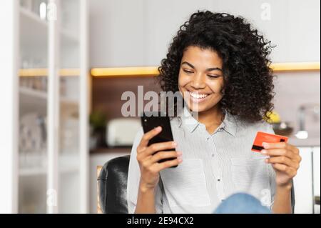 Jeune fille afro-américaine souriante avec des cheveux bouclés tenant le téléphone mobile, en entrant le numéro de carte de crédit pour faire une transaction en ligne, race mixte Banque D'Images