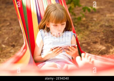 Portrait d'une petite fille excitée regardant l'écran du téléphone mobile, allongé à l'extérieur sur un hamac dans le parc Banque D'Images