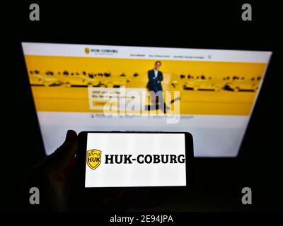 Personne portant un smartphone avec le logo de la compagnie d'assurance allemande HUK-Coburg exposé devant le site Web d'affaires. Mise au point sur l'écran du téléphone. Banque D'Images