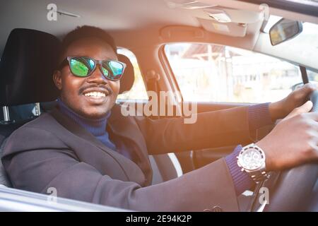 Homme africain en costume noir assis derrière le volant avec sourire et heureux Banque D'Images