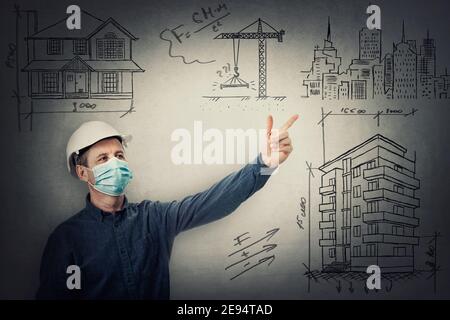 Entrepreneur d'âge moyen ou architecte portant un casque de protection et un masque facial comme prévention Covid-19. Ingénieur avec hardhat pointant l'index showin Banque D'Images