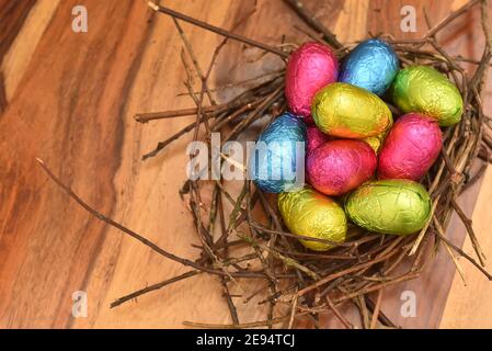 Des œufs de pâques colorés enveloppés de feuilles dans du rose, du vert, du bleu et du jaune dans un nid naturel fait de bâtons et de brindilles, contre un dos en bois brun multigrain Banque D'Images