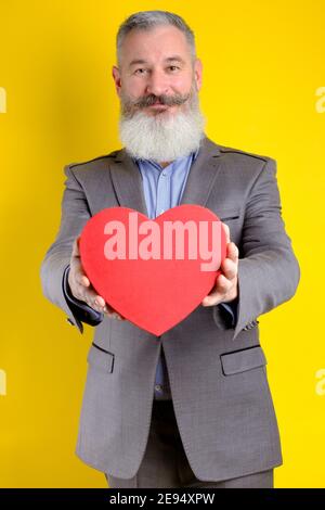 Studio portrait d'homme barbu mature tenant une boîte cadeau en forme de coeur, fond jaune, espace de copie Banque D'Images
