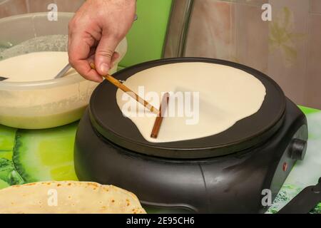 Cuisine du petit déjeuner dans une crêpe électrique. La main de l'homme verse la pâte et la répand sur la surface avec le bâton. Mise au point sélective, gros plan. Banque D'Images