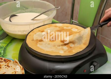 Faire cuire des crêpes à la maison dans une poêle électrique ronde. Le chef retourne la pâtisserie avec une spatule. Mise au point sélective, gros plan. Banque D'Images