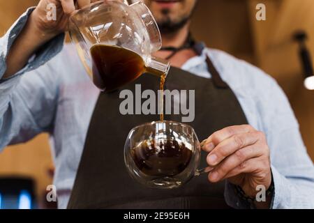 Gros plan versez du café dans une tasse en verre double dans un café par un beau barista barbu. Autres méthodes de préparation du café : syphon et aéropress. Publicité pour s Banque D'Images