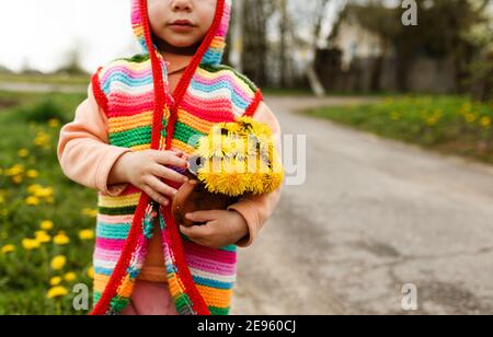 une petite fille dans une veste multicolore lumineuse se tient dans la rue avec un bouquet de pissenlits jaunes. Banque D'Images