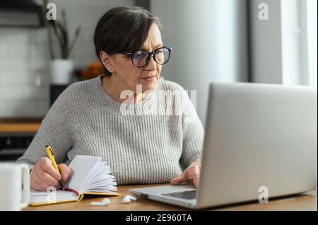 Femme en retraite concentrée utilisant un ordinateur portable assis au bureau dans la cuisine pour travailler à distance, étudier en ligne, regarder un webinaire et prendre des notes Banque D'Images