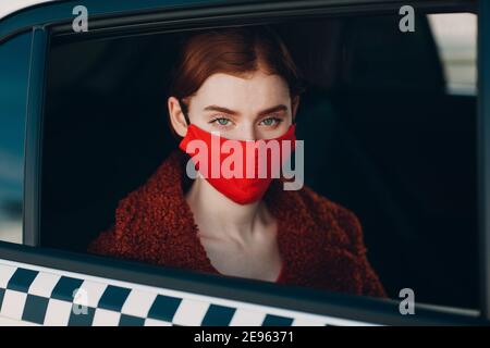 Une jeune femme au gingembre prend un trajet en voiture de taxi pendant la quarantaine pandémique du coronavirus. Femme portant un masque médical rouge stérile. Concept de la distance sociale et de la sécurité sanitaire dans les transports Banque D'Images