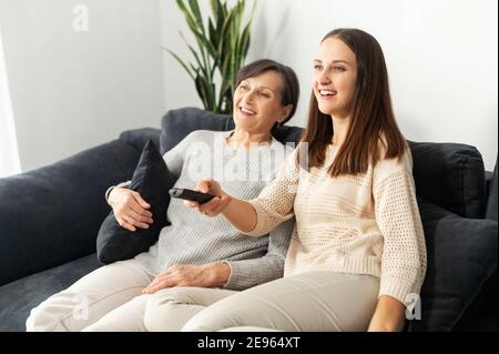 Une mère âgée et une fille adulte se détendent sur un canapé à la maison, regardant la télévision, changeant de chaîne avec télécommande, souriant et appréciant le temps Banque D'Images