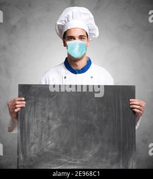 Chef masqué ou cuisinier tenant un tableau noir pendant la pandémie du coronavirus covid, restaurants et concept alimentaire Banque D'Images