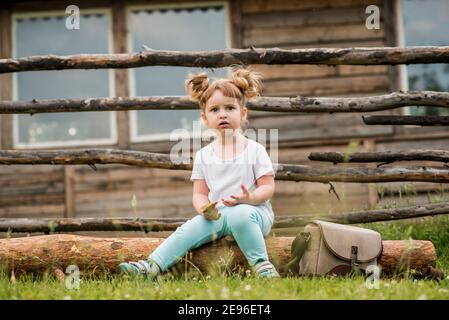 Portrait extérieur d'une fille assise sur l'herbe près de la clôture.été dans le village. Belle fille de bébé sur un banc de bois.écologie et heureux Banque D'Images