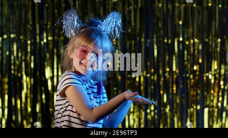 Mignon enfant regarder l'appareil photo dans la boîte de nuit présentant le produit, le logo ou le texte. Jeune fille ado élégante debout sur des lumières néon bleues et des points à l'espace vide, signe d'accord, se tromper autour. Espace-copie Banque D'Images