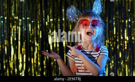 Adolescente élégante fille debout sur des lumières néon bleu rétroéclairées et des points à l'espace vide, signe d'accord, se tromper autour. Joli enfant regarde l'appareil photo dans la boîte de nuit. Présenter le produit, le logo ou le texte. Espace-copie Banque D'Images