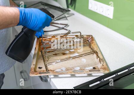 un homme portant des gants en caoutchouc bleu nettoie les grilles sales et plaques de cuisson du four avec un spray nettoyant Banque D'Images