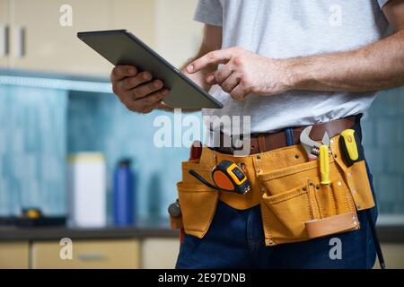Coupe courte d'un jeune réparateur portant une ceinture à outils avec divers outils utilisant une tablette numérique en intérieur Banque D'Images