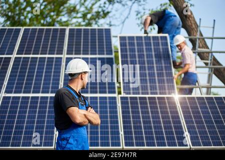 Profil du jeune ingénieur technicien debout devant le panneau solaire extérieur haut non fini. Système photovoltaïque bleu surface brillante observant l'équipe de travailleurs sur une plate-forme en acier haute. Banque D'Images