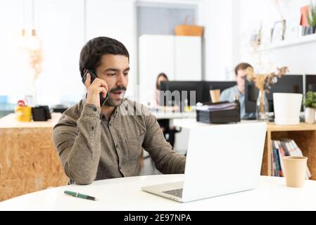 Décontracté habillé hispanique homme parlant sur le téléphone devant son ordinateur portable dans le bureau. Banque D'Images