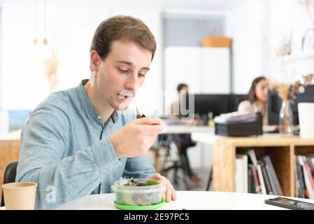 Concept d'alimentation saine en milieu de travail. Beau travailleur ayant une pause déjeuner et en mangeant une salade dans le bureau. Banque D'Images