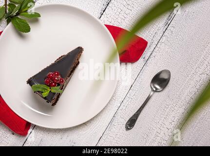 Morceau de gâteau au chocolat appelé Mozart décoré de baies vitrées de cassis rouge et de feuille de menthe sur une assiette blanche avec une cuillère, une nappe rouge et sur une W blanche Banque D'Images