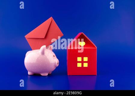 Enveloppe rouge festive insérée dans une tirelire rose à côté d'une petite maison rouge de papier avec chaud lumière dans les fenêtres sur fond bleu avec cop Banque D'Images