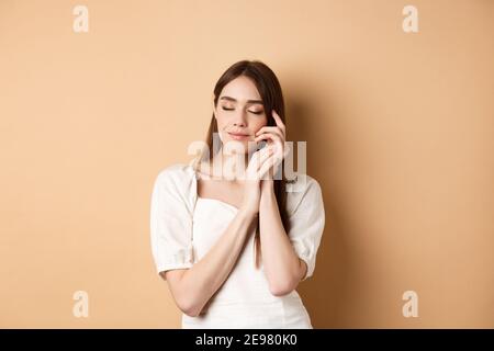 Femme tendre et romantique proche des yeux, touchant doucement la peau douce et lisse, souriant rêveur tout en se tenant sur fond beige Banque D'Images