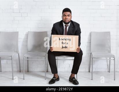 Homme afro-américain en costume formel tenant À LA RECHERCHE de L'AFFICHE D'EMPLOI, en attente de l'entretien de travail dans le hall de bureau, espace libre Banque D'Images