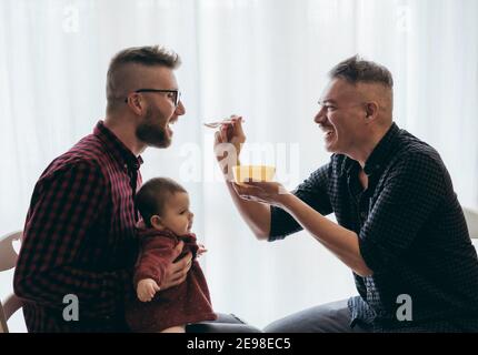 Homme gay couple avec adopté bébé fille à la maison - Deux beaux pères nourrissent la petite fille dans la cuisine - Baby-sitters masculins - famille LGBT à la maison - diversité Banque D'Images