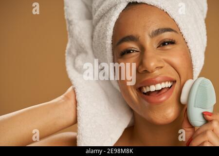 Gros plan de la jeune femme joyeuse après la douche regardant excitée de l'appareil photo tout en utilisant la brosse de nettoyage du visage en silicone, posant isolé sur fond beige. Beauté, concept de soins spa Banque D'Images