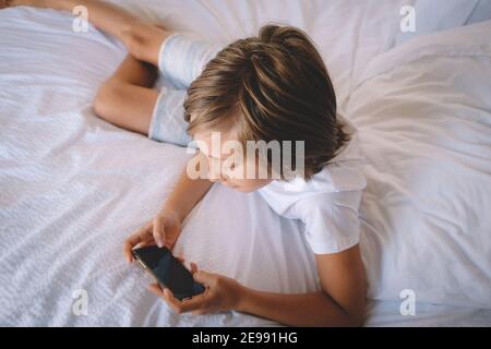 Boy in White vérifie sur son téléphone depuis un lit de chambre d'hôtel. Banque D'Images