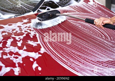 Voiture rouge lavée dans un lave-auto libre-service sur la brosse se déplaçant sur la capuche avant, laissant des coups dans le savon blanc Banque D'Images