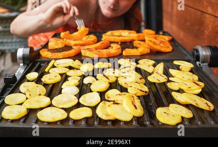 Chips de pommes de terre et morceaux de courge de noyer cendré grillés sur le gril électrique, se concentrer sur les cercles de pommes de terre brillants, flou jeune femme en arrière-plan Banque D'Images