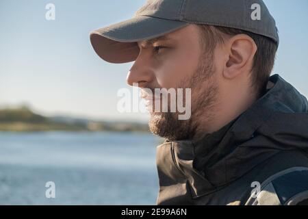 Vue latérale d'un homme barbu en toute confiance dans un casquette de baseball regardant la distance sur l'arrière-plan de un lac ou une rivière par une journée ensoleillée en automne Banque D'Images