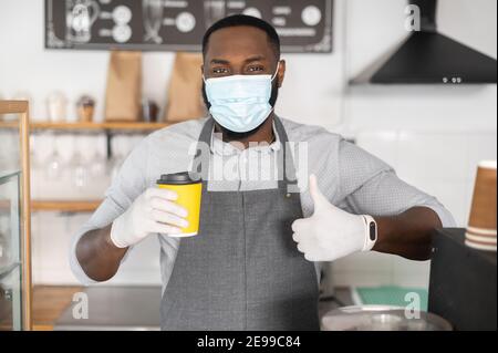 Portrait en gros plan serveur afro-américain masculin, le barista portant un masque tient une tasse de café en papier et montre le pouce debout derrière le comptoir dans un cadre moderne Banque D'Images