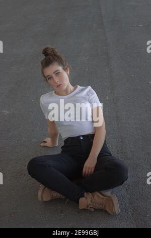 Magnifique femme brune aux yeux bleus dans un petit pain regardant un appareil photo avec une expression calme, assis et penchée dans la rue. Modèle de mode extérieur 2021. Banque D'Images