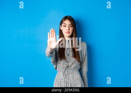 Femme sérieuse et confiante avec de longs cheveux et de la robe, étirez la main et dites non, faites un geste d'arrêt, interdisant les mauvaises actions, debout sur fond bleu Banque D'Images