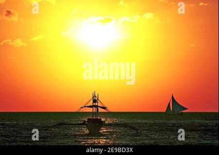 Un bateau banca sur l'eau est souligné par le coucher de soleil orange et jaune au large de la côte de l'île Boracay aux Philippines. Banque D'Images