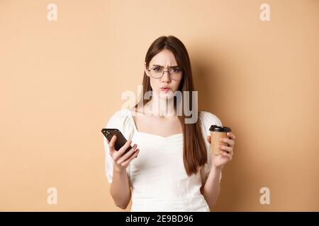 Une fille confuse dans des verres frognant à l'appareil photo après avoir lu un téléphone portable, tenant une tasse de café, debout déplaisée sur fond beige Banque D'Images