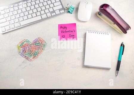 Mots Happy Valentine's Day dans une note sur le bureau avec des trombones de couleur en forme de coeur et de clavier, stylo, bloc-notes et agrafeuse à la station de travail Banque D'Images