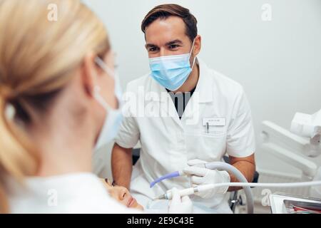 Dentiste de sexe masculin discutant avec une assistante de sexe féminin lors du traitement d'un patient. Les médecins dentaires parlent du traitement de la patiente assise à denti Banque D'Images