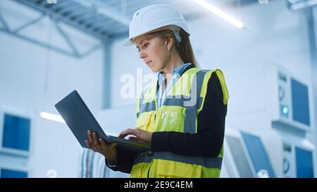 Usine de haute technologie : femme ingénieure confiante et professionnelle portant une veste de sécurité et un casque de sécurité tenant et travaillant sur un ordinateur portable. Moderne et lumineux Banque D'Images