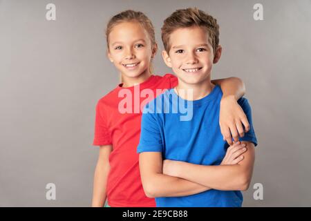 Adorable femme enfant enroulant le bras autour de l'épaule de frère et souriant pendant que le garçon croise les bras au-dessus de sa poitrine Banque D'Images