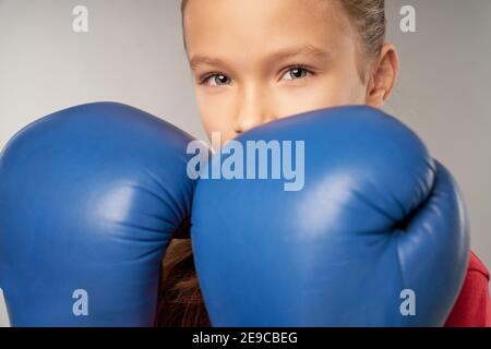 Gros plan du boxeur féminin pour enfants couvrant le visage avec les mains dans des gants de boxe tout en regardant la caméra et en faisant des combats position Banque D'Images