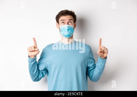 Coronavirus, concept de santé et de quarantaine. Curieux gars lisant la bannière sur le dessus, regardant et pointant les doigts vers le haut, debout dans le masque médical sur blanc Banque D'Images
